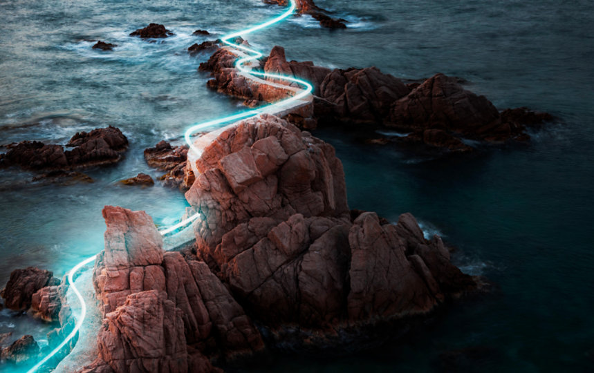 A line of blue light painted along a foot path winding through dramatic red rock formations on the Costa Brava coastline.